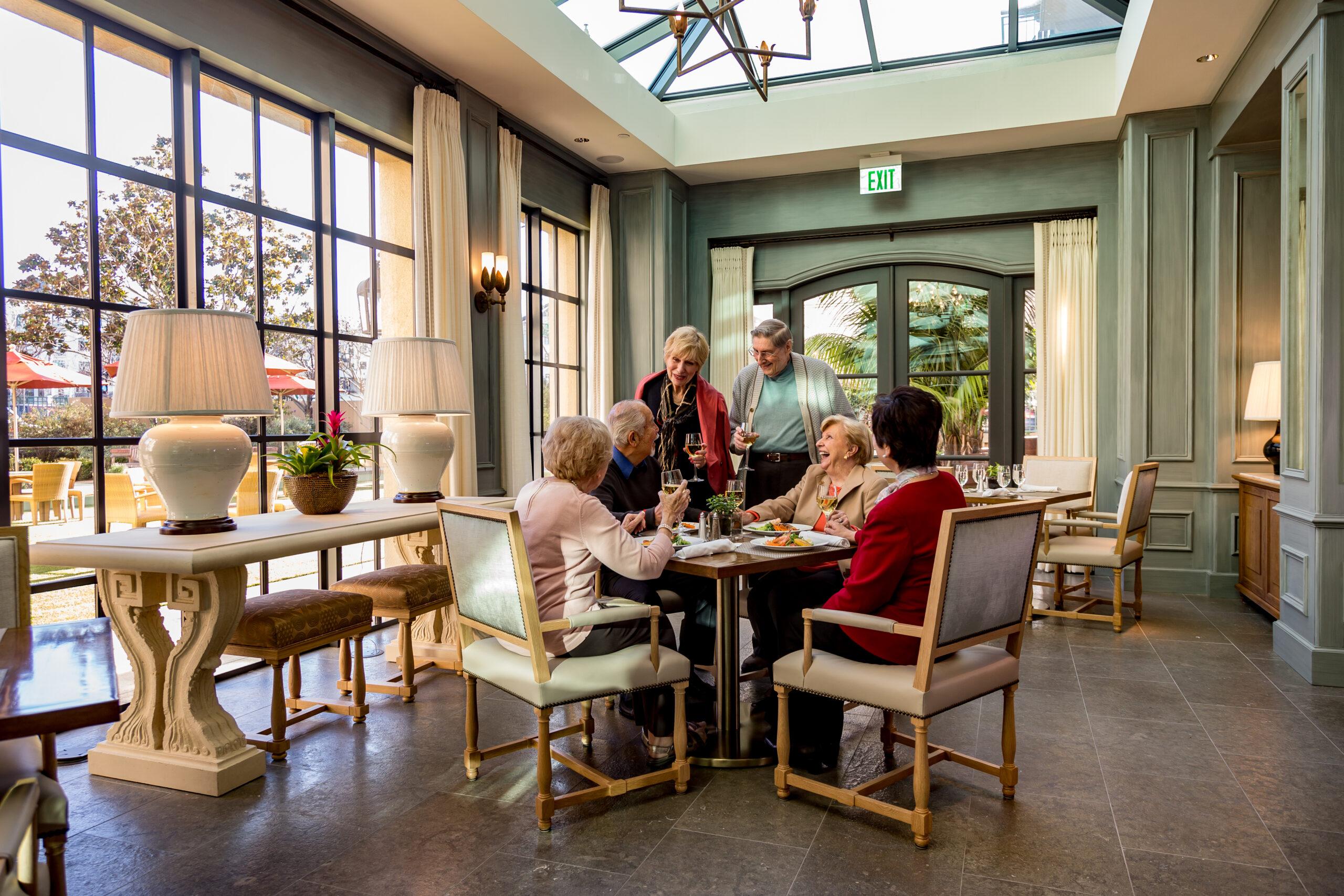 Older adults talk and laugh in the sun-filled atrium dining room