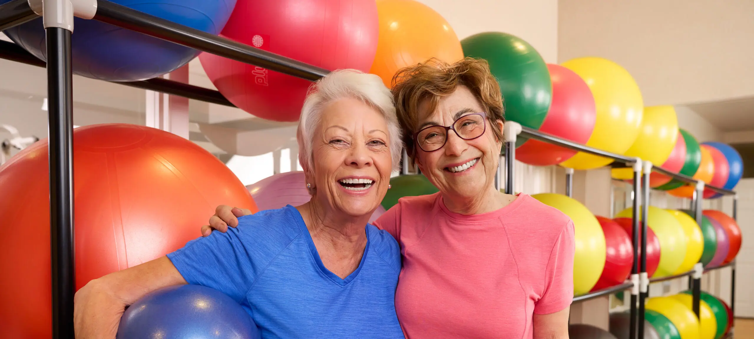 Two women smile for the camera, holding exercise equipment, in front of colorful exercise balls.